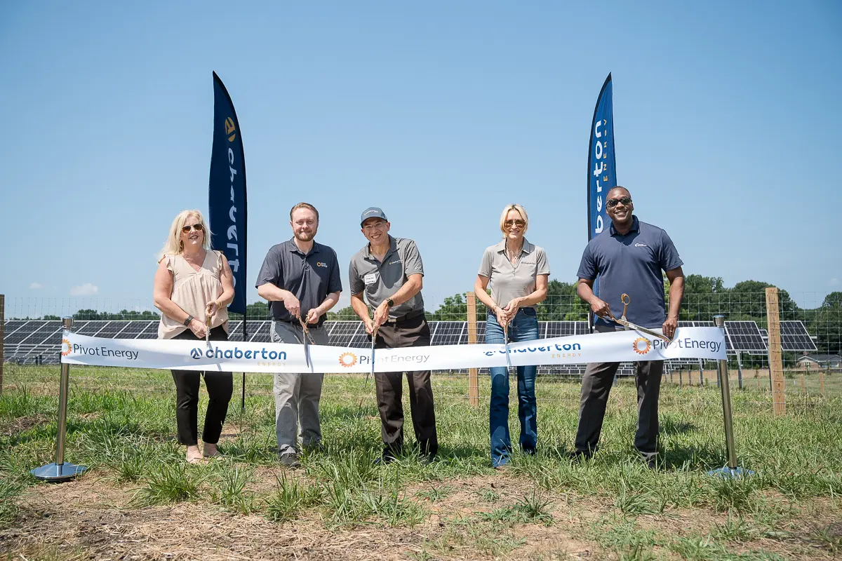 Five executives stand in front of a solar farm about to cut a ceremonial ribbon