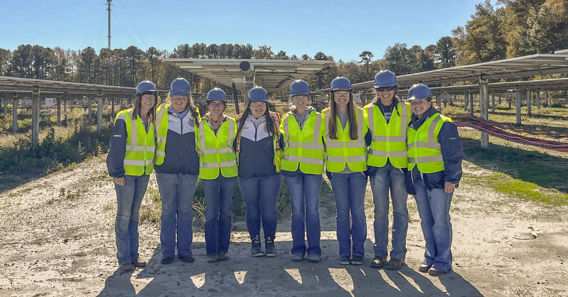 Eight female high school students wear hard hats and safety vests while smiling at the camera at a solar development site